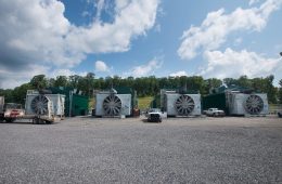 Four large box-like structures with huge fans sits across a gravel lot