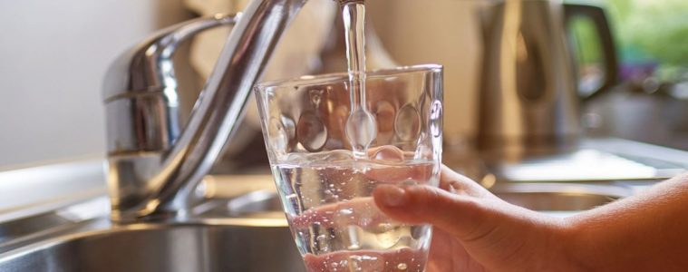 A hand holds a glass in a kitchen sink that is being filled with water