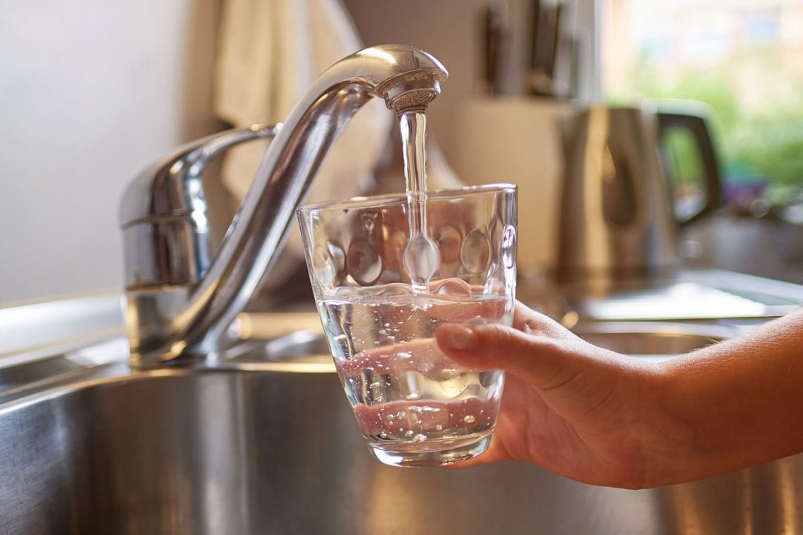 A hand holds a glass in a kitchen sink that is being filled with water