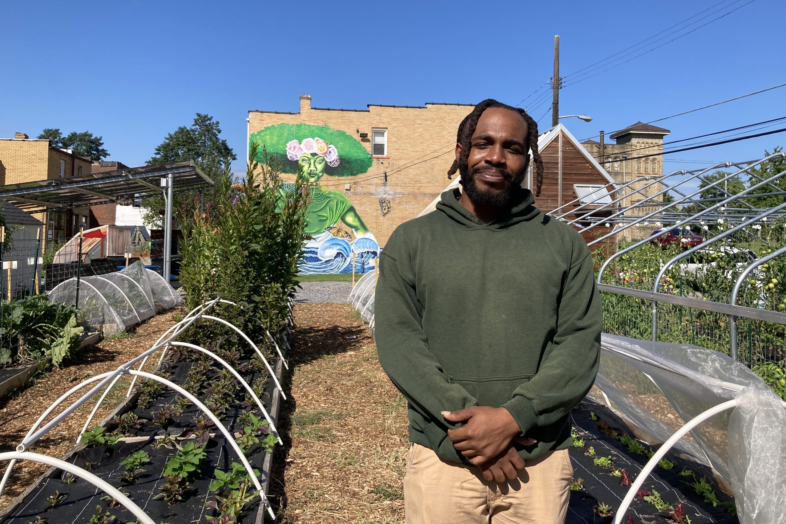 Tacumba Turner in front of covered rows of vegetables, and a building with a mural