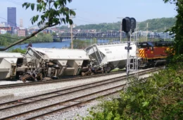 derailed train cars along tracks near Mon River with Downtown Pittsburgh in background