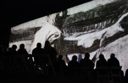 Silhouette of people from behind, seated, watching a black and while image of a worker projected on a wall