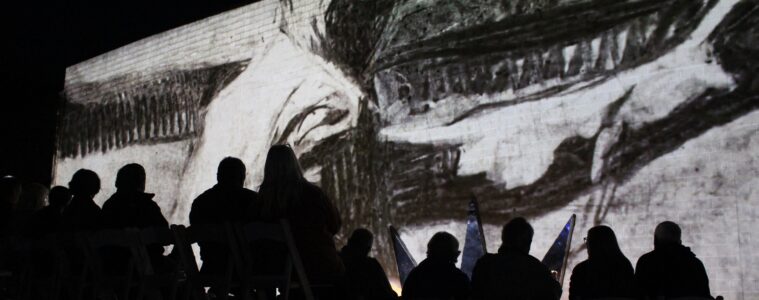 Silhouette of people from behind, seated, watching a black and while image of a worker projected on a wall