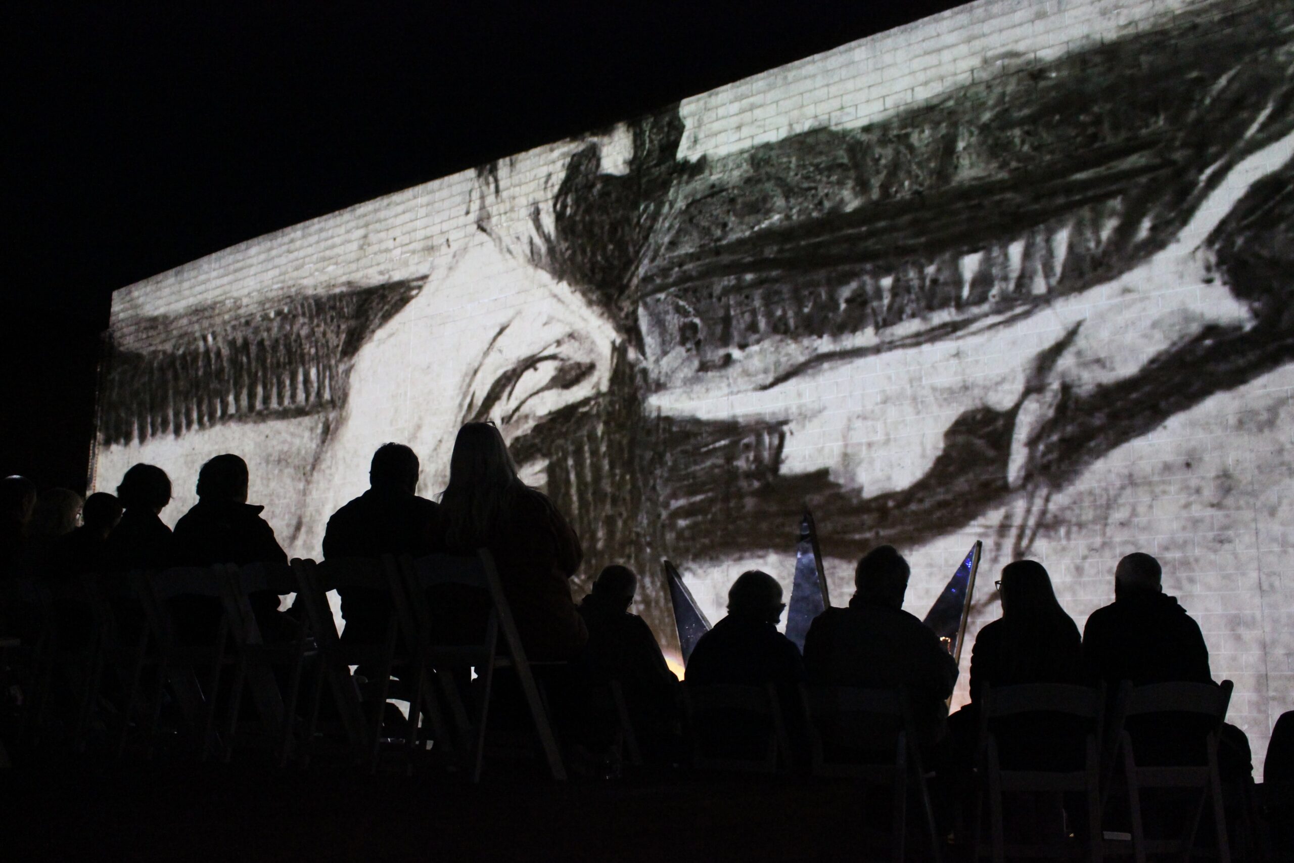 Silhouette of people from behind, seated, watching a black and while image of a worker projected on a wall