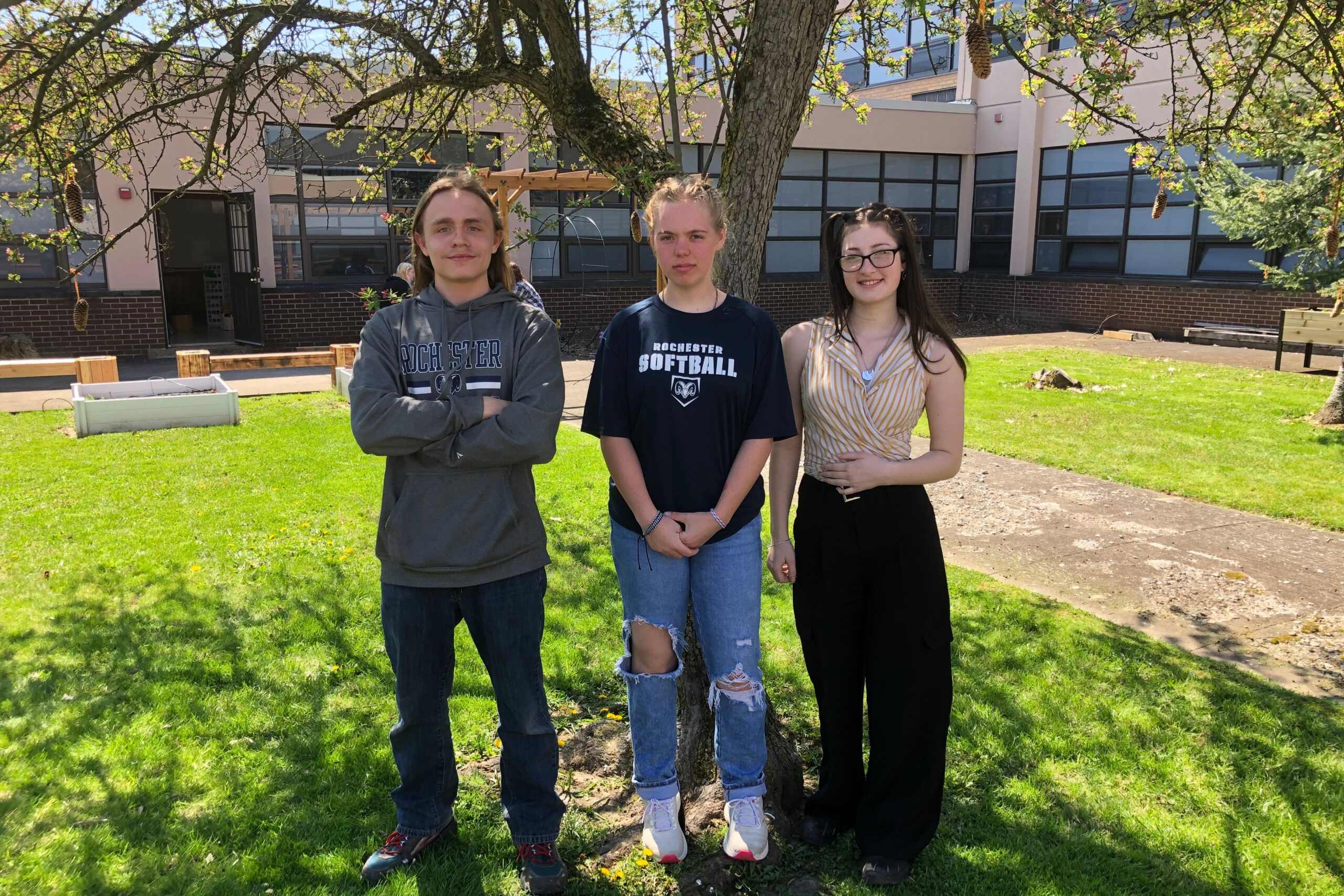 Three students stand under a tree on the school grounds