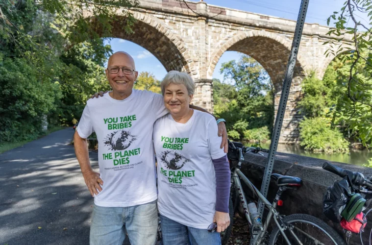Two riders, arm in arm, stand on a road next to their bike, with a stone overpass behind them