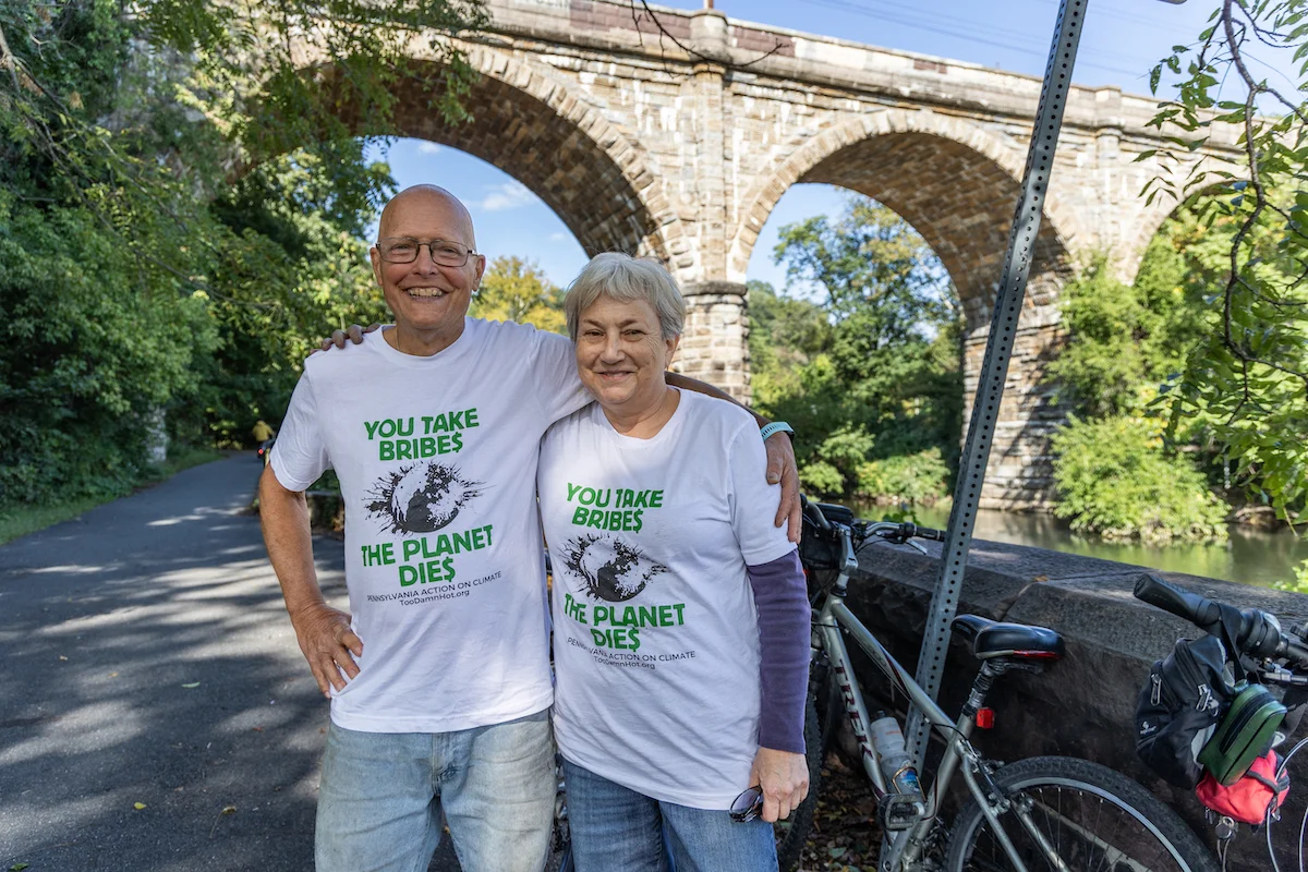 Two riders, arm in arm, stand on a road next to their bike, with a stone overpass behind them