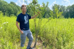 Chris Kubiak stands next to an apple tree that is a little taller than him, surrounded by tall wildflowers.