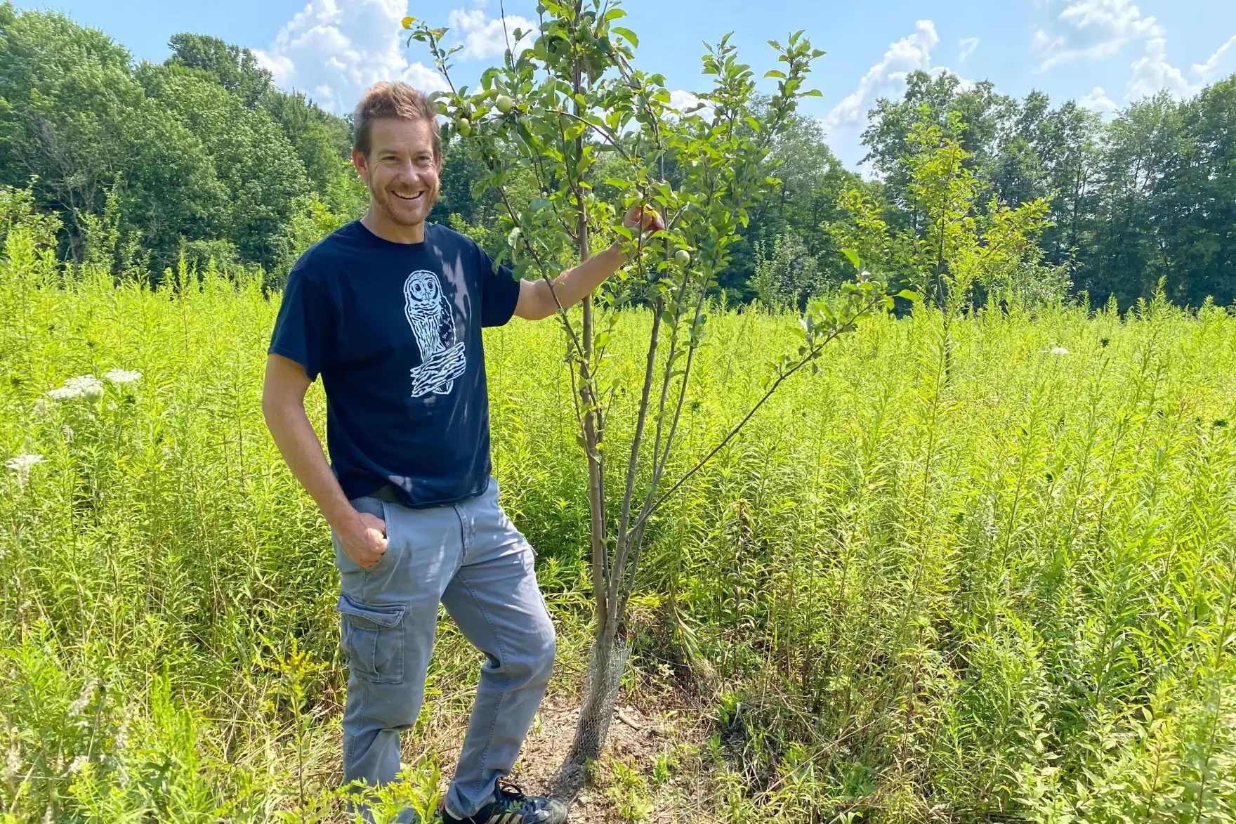 Chris Kubiak stands next to an apple tree that is a little taller than him, surrounded by tall wildflowers.