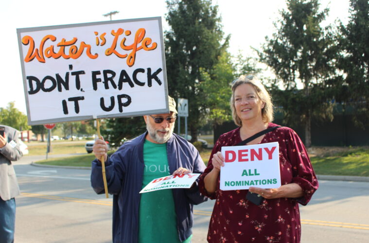 One protestor holds a sign reading "Water is life, don't frack it up." A woman standing next to him holds a sign that reads, "Deny all nominations."