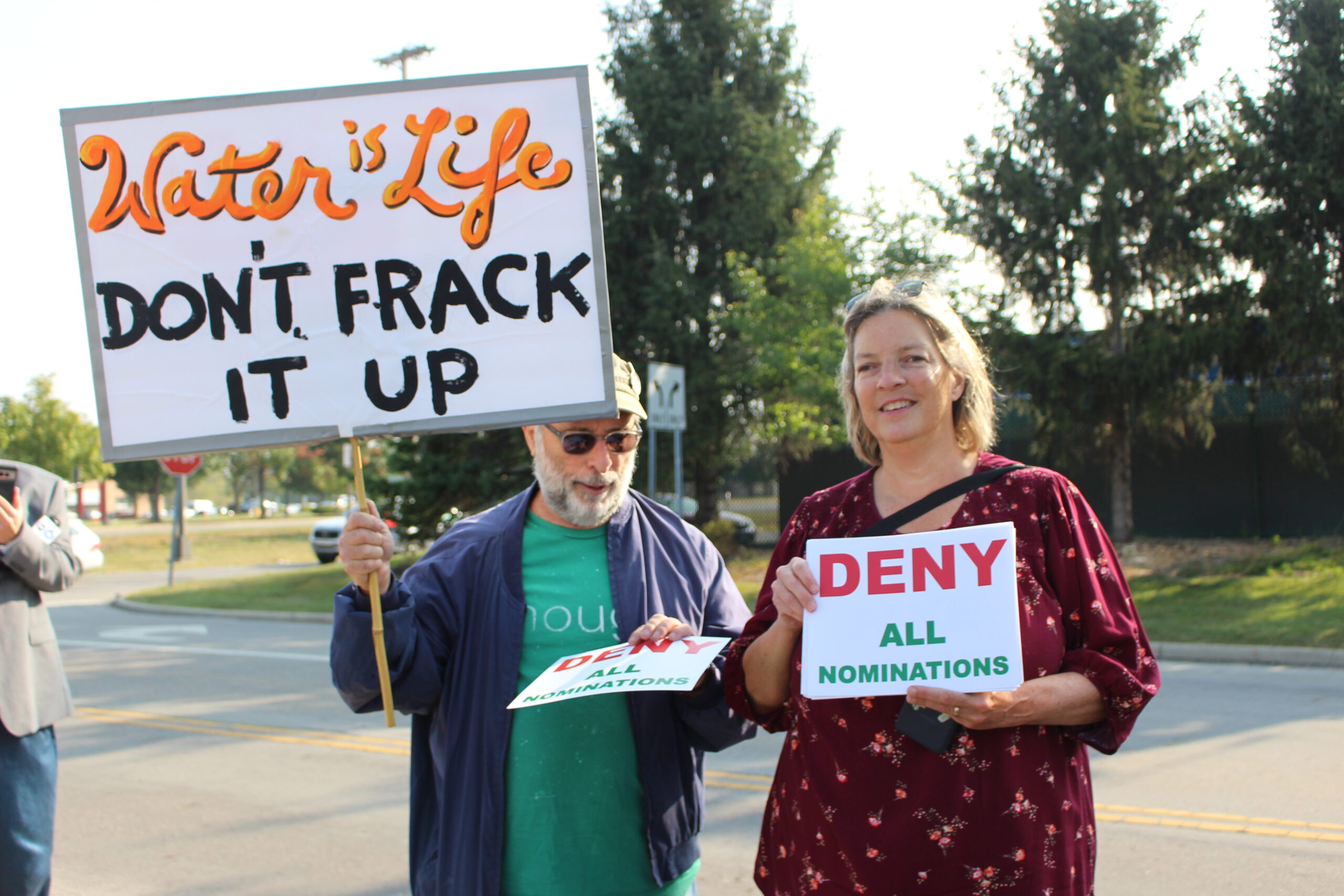 One protestor holds a sign reading "Water is life, don't frack it up." A woman standing next to him holds a sign that reads, "Deny all nominations."