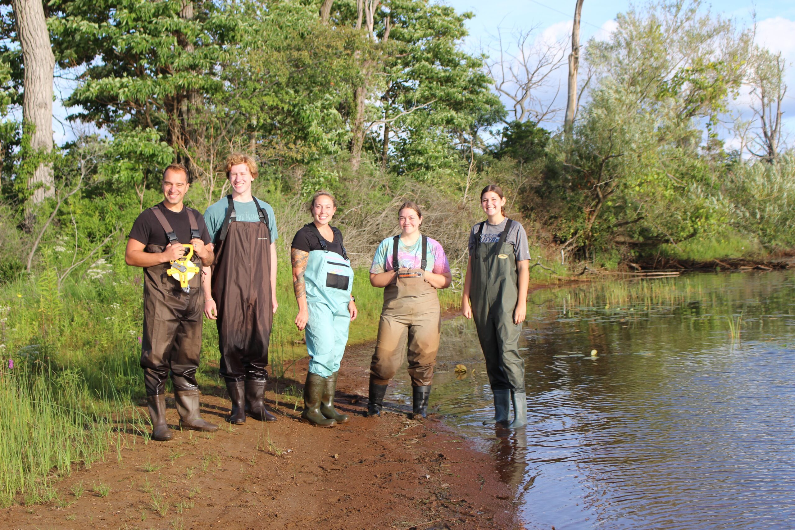 5 people in waders stand at the edge of a lake