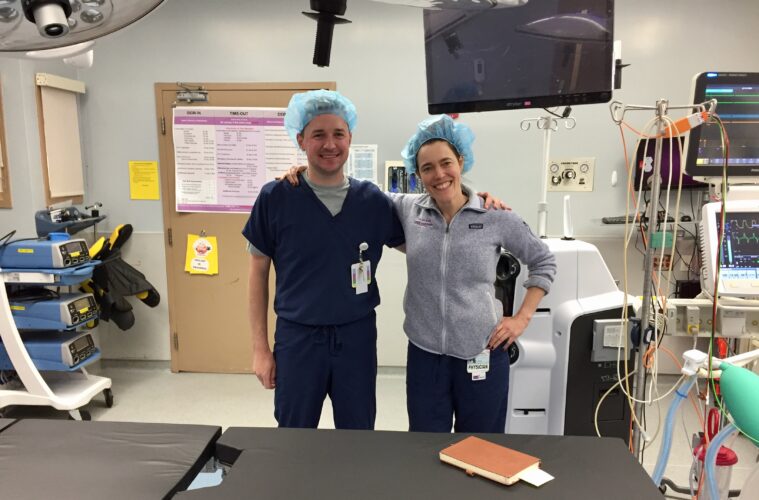 Pete Adams and Noe Woods stand arm in arm in an operating room wearing scrubs
