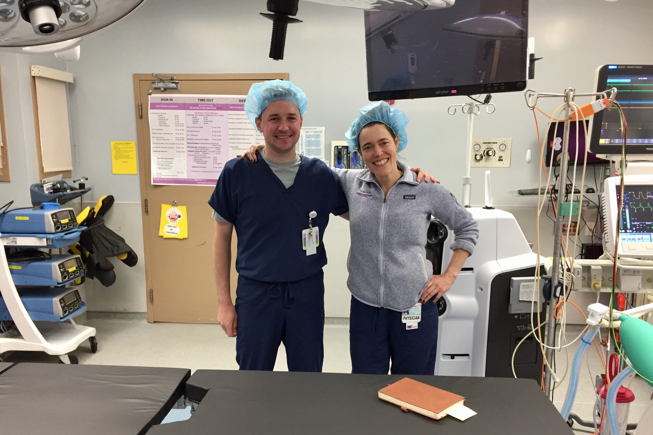 Pete Adams and Noe Woods stand arm in arm in an operating room wearing scrubs