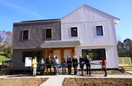 Eleven people state in front of a duplex holding a large ceremonial ribbon