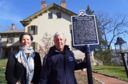 Mary Sorensen in a coat with a white scarf stands to the left of Roger Williams in a blue "Penn State Alumni" jacket next to a blue historical marker, with an historic house in the background
