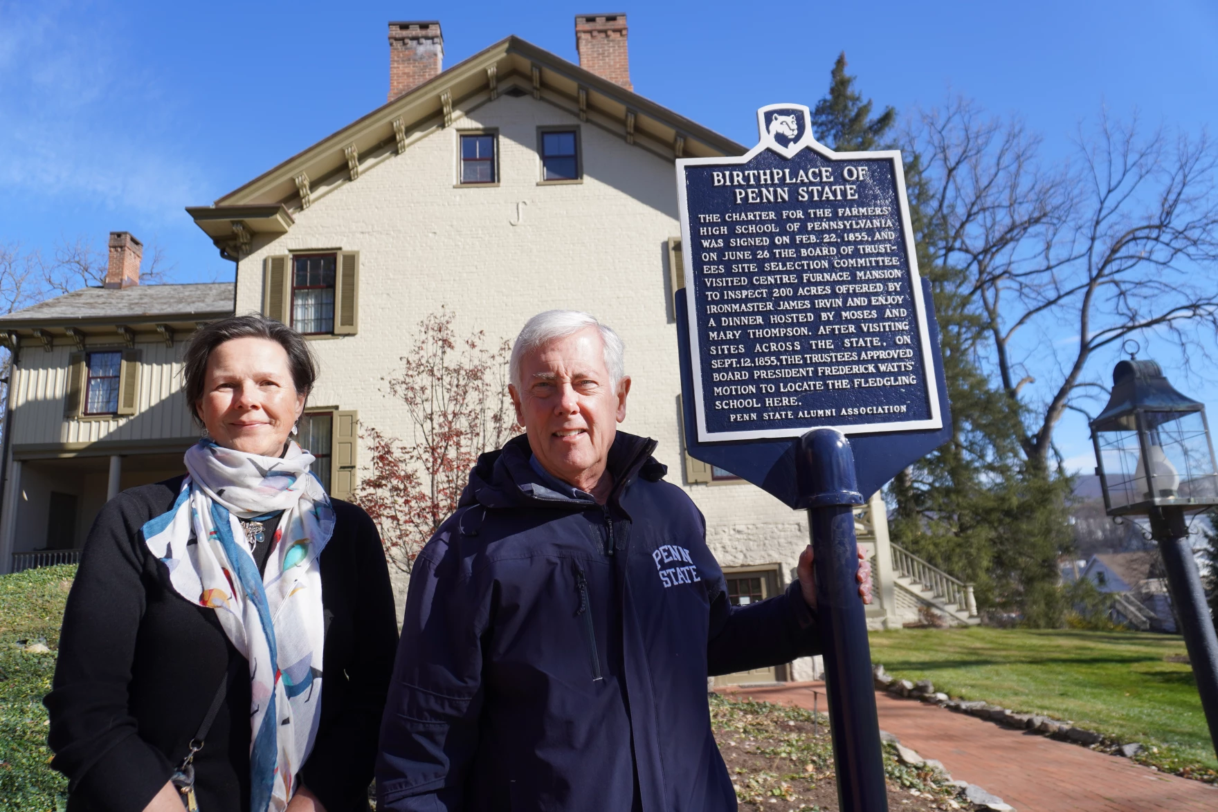 Mary Sorensen in a coat with a white scarf stands to the left of Roger Williams in a blue "Penn State Alumni" jacket next to a blue historical marker, with an historic house in the background