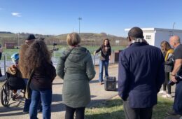 A woman with a microphone talks to a group of people surrouning her against a blue sky.