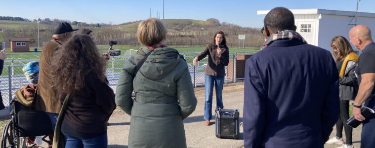 A woman with a microphone talks to a group of people surrouning her against a blue sky.
