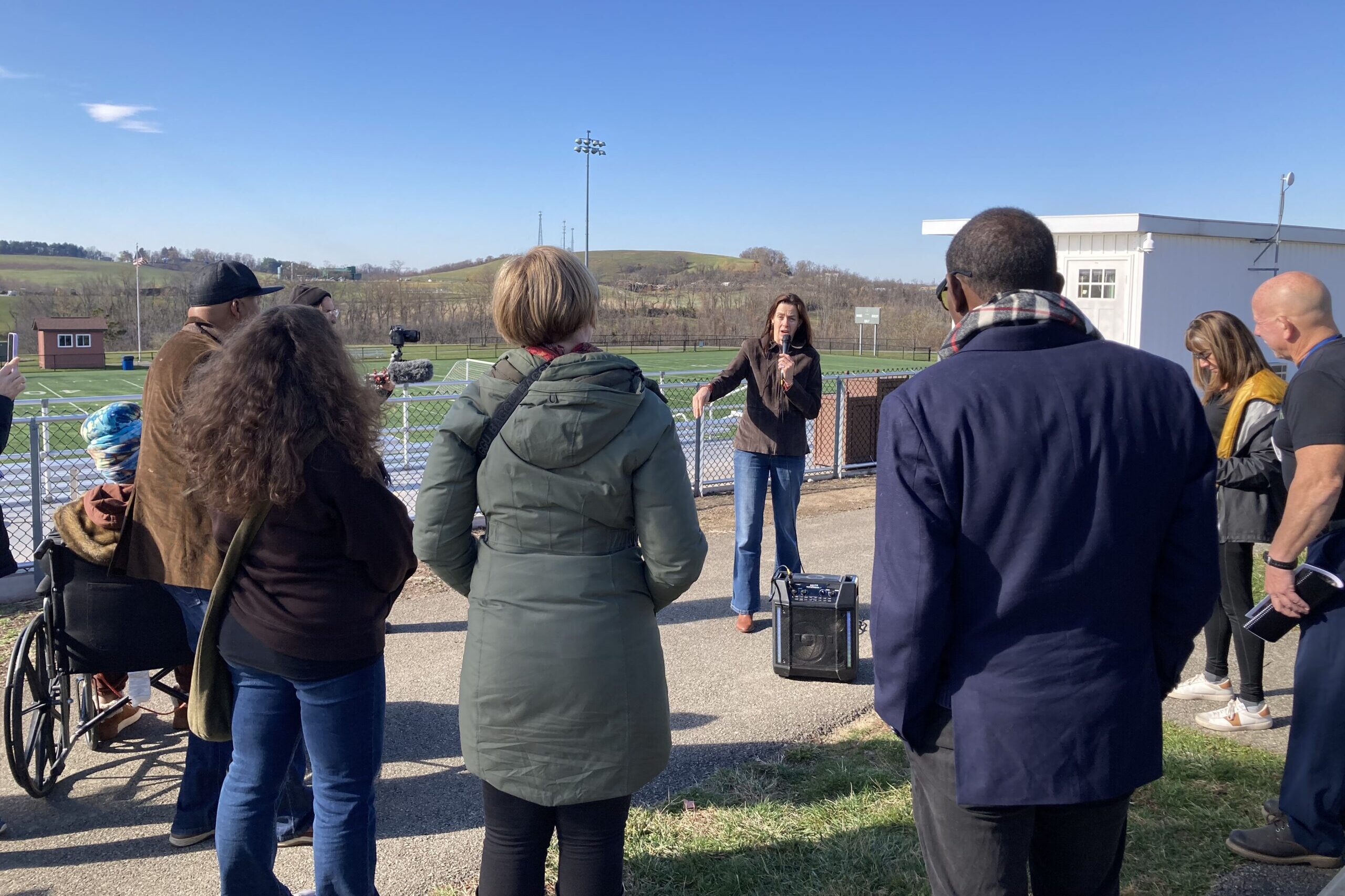 A woman with a microphone talks to a group of people surrouning her against a blue sky.