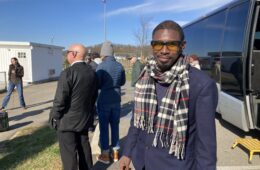 A Black man with a plaid scarf, sunglasses and blue coat looks at the camera as a group of people are listening to a speaker behind him, with a tour bus on the right