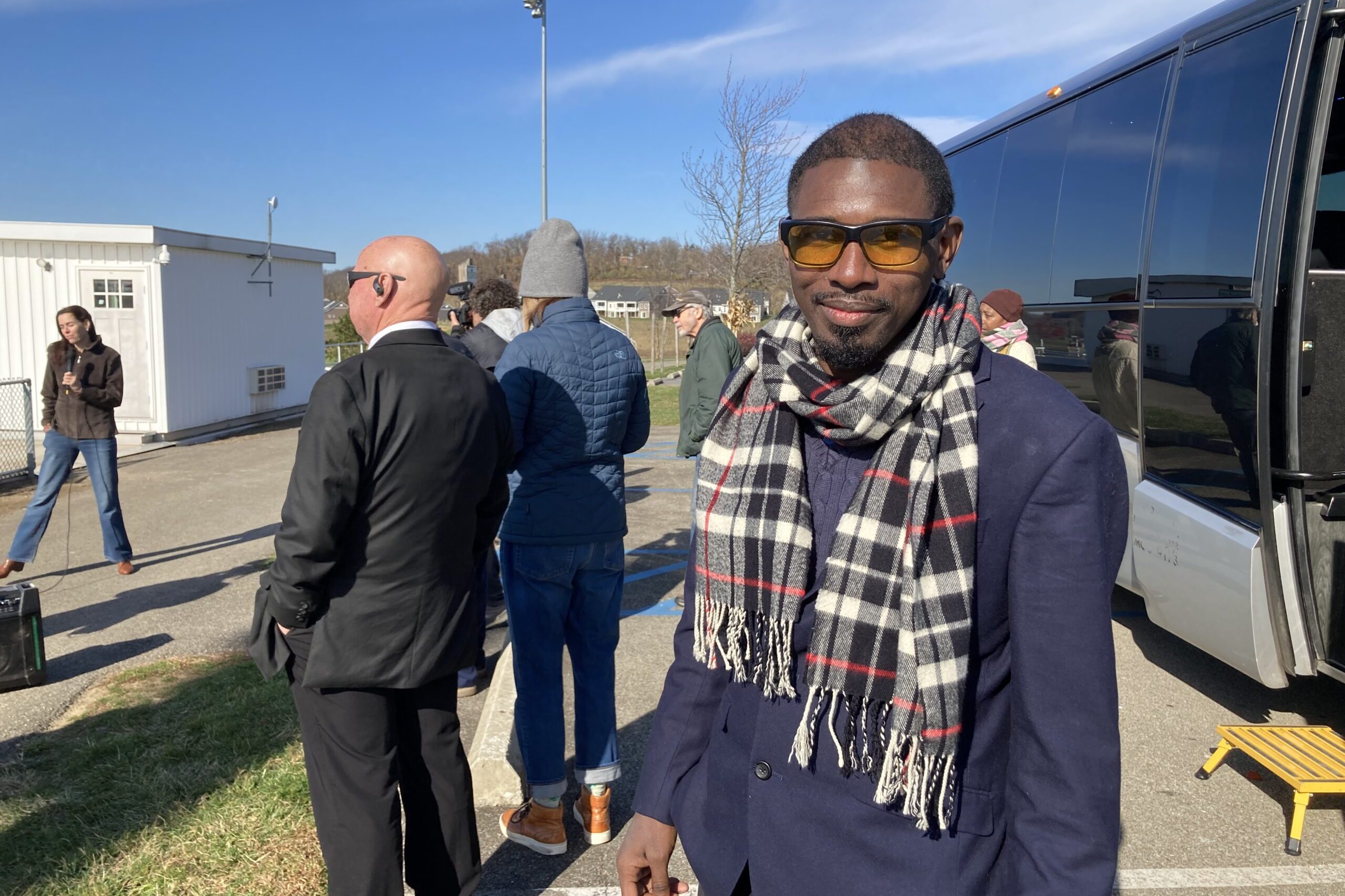 A Black man with a plaid scarf, sunglasses and blue coat looks at the camera as a group of people are listening to a speaker behind him, with a tour bus on the right
