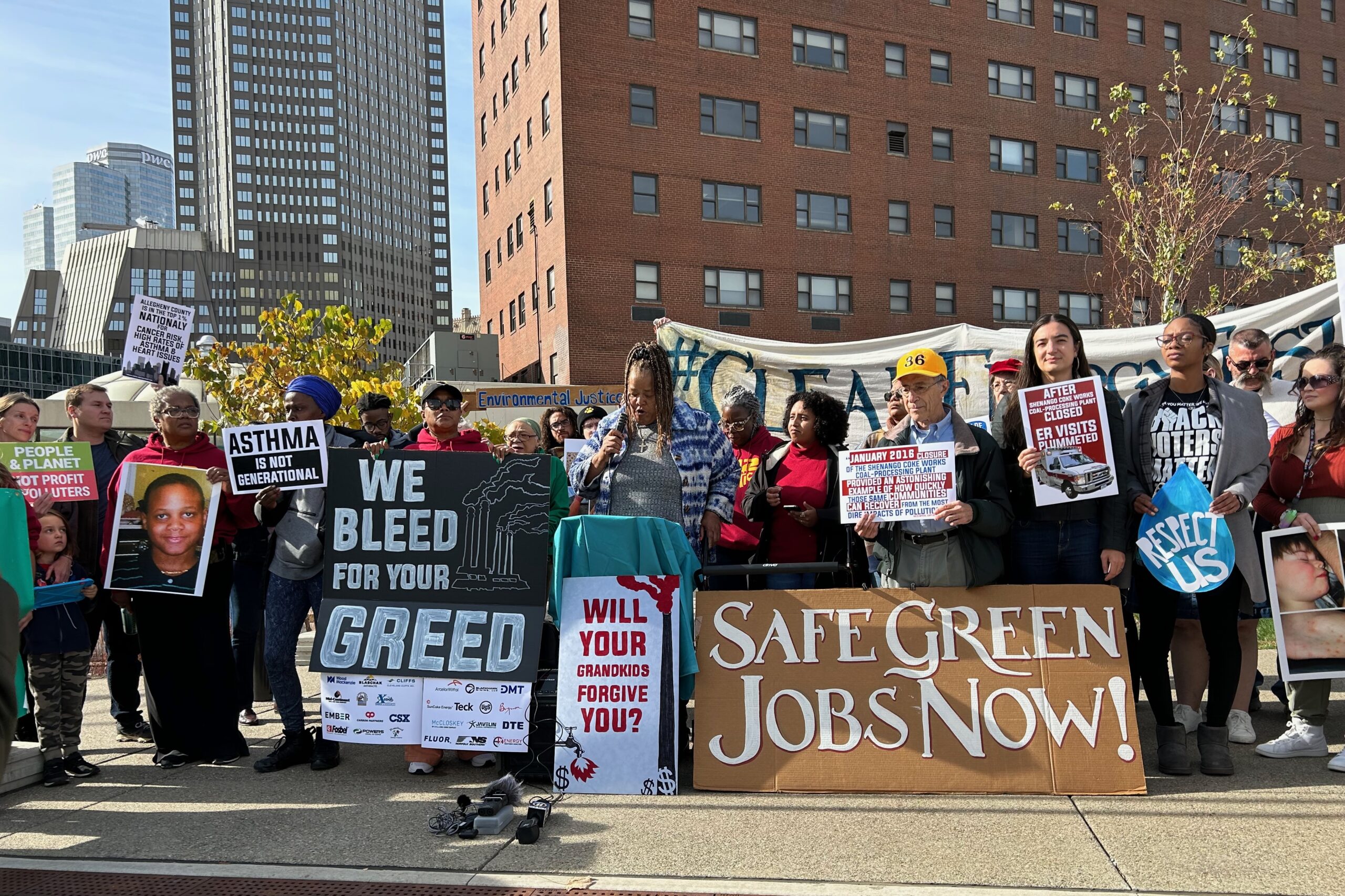 Protestors stand behind signs that read "Safe clean jobs now" and "We bleed for your greed," among others. A Black woman stands in the middle, speaking into a microphone