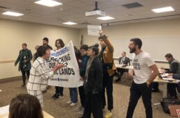 A woman in white and black outfit shouts at protestors standing feet away holding a sign and shouting back, in a beige conference room.