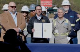 Governor Shapiro stands in front of a table holding a signed agreement with 2 men on either side of him.