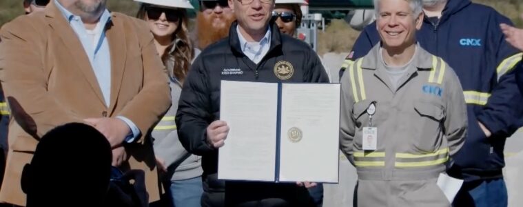 Governor Shapiro stands in front of a table holding a signed agreement with 2 men on either side of him.