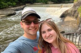 A man in a baseball hat and sunglasses, left, with a woman in a pink shirt with a concrete waterfall in the background.