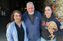 A man in a blue sweatshirt stands between two women. One is holding a small dog.