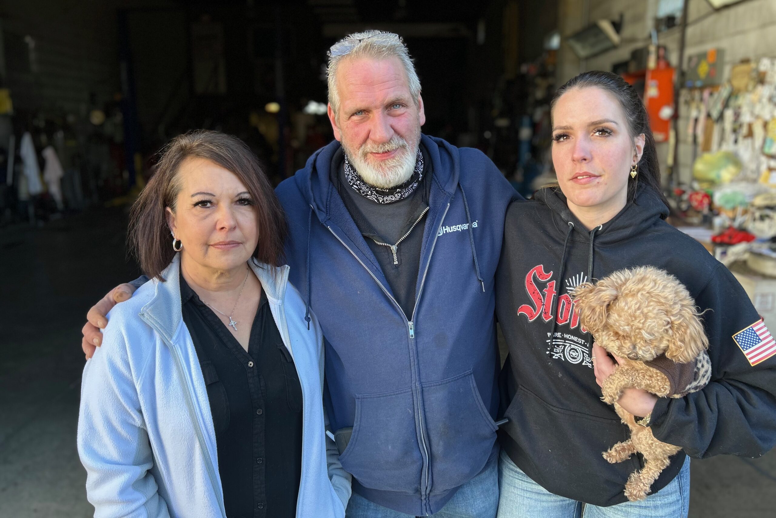 A man in a blue sweatshirt stands between two women. One is holding a small dog.