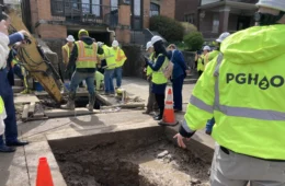 Workers in bright yellow jackets reading PGH2O standing around 2 holes cut in the road where water lines run