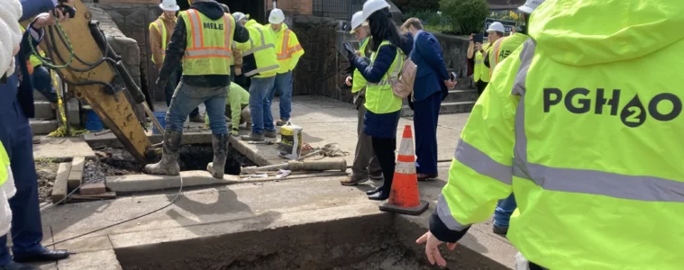 Workers in bright yellow jackets reading PGH2O standing around 2 holes cut in the road where water lines run