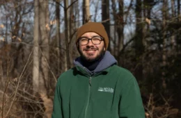A man with a brown knit hat and green hoodie with glasses and a beard stands in a forest