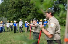 Two instructors wearing DCNR uniforms holding red poles stand in front of a group of hard-hat wearing young adults.