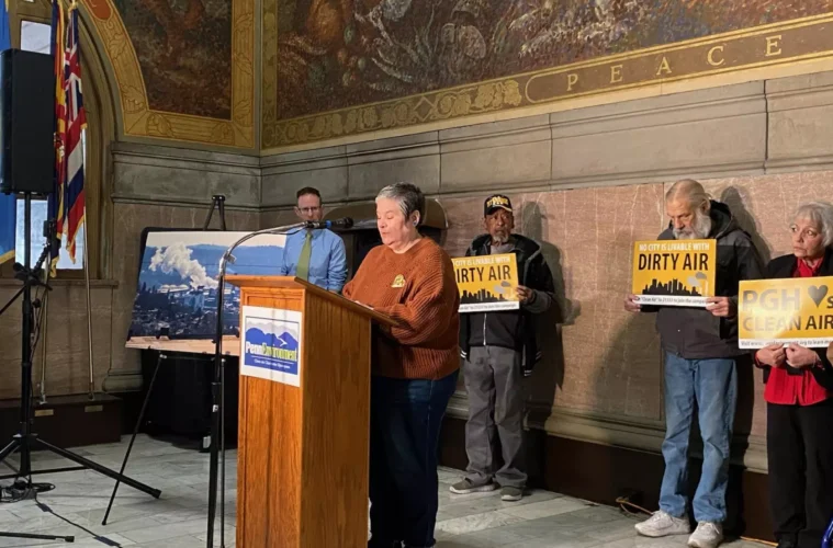 A person stands at a podium, while 3 others line up behind her holding signs that read "Dirty Air."