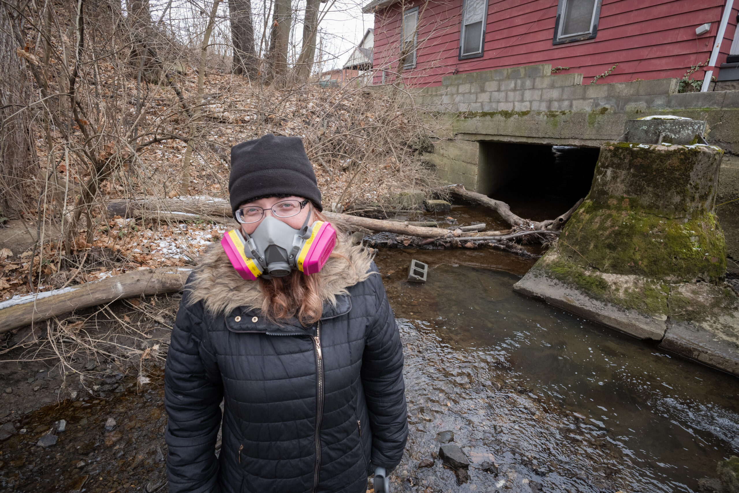 A woman stands in a stream in front of a culvert going under a home. She is wearing a winter coat and hat with a respirator.