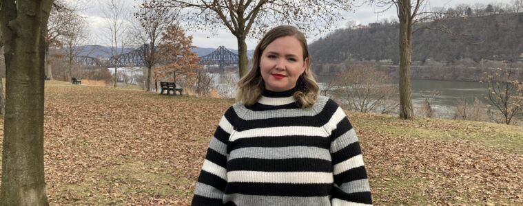 A young woman in a black sriped sweater stands in front of a river in a park.