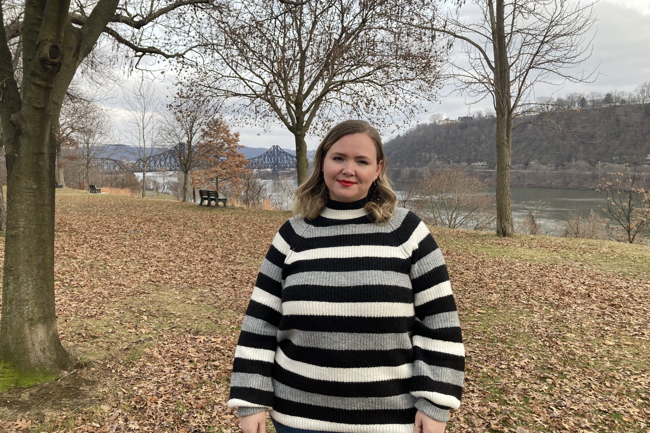 A young woman in a black sriped sweater stands in front of a river in a park.