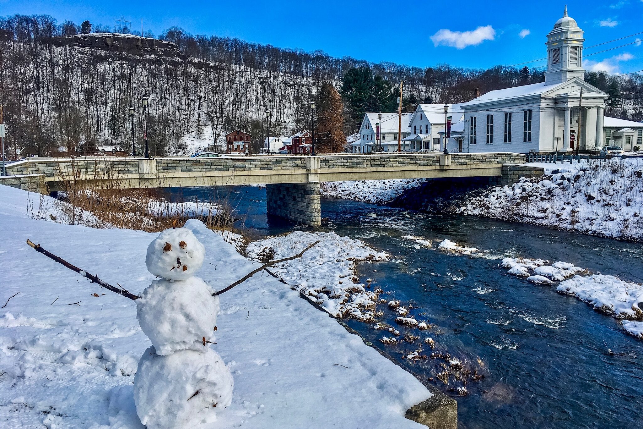 The Lachawaxen River with a bridge and a church in the background, and a snowman in the foreground