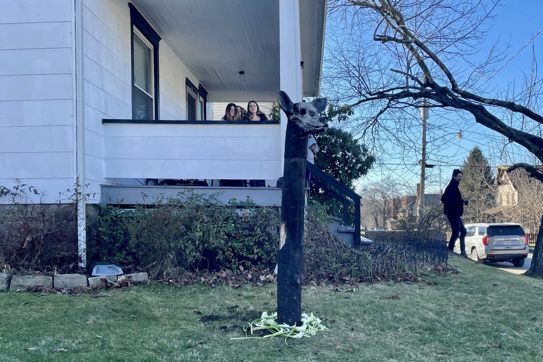 A deer head is carved on a burnt log stands in the side yard of a white clapboard house. It is faced looking toward the right, across the street.