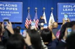 Kamala Harris in front of a podium with flags and blue signage behind her, with a room full of people in the foreground.