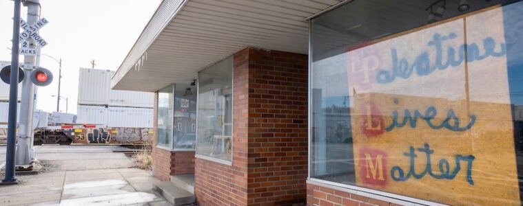 A storefront with a handmade yellow sign that reads "Palestine lives matter" with a train crossing signal across the street.