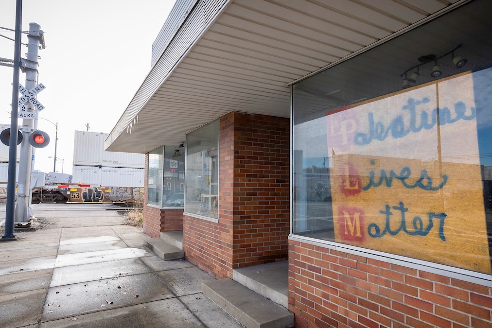 A storefront with a handmade yellow sign that reads "Palestine lives matter" with a train crossing signal across the street.