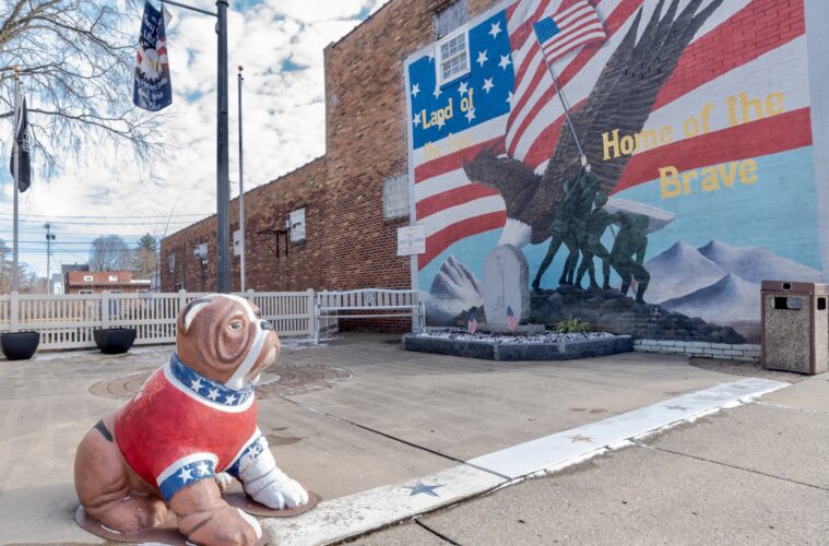 A statue of a bulldog painted red, white and blue sits along the sidewalk with a patriotic mural in the background
