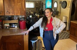 A woman in a red shirt and tan cardigan and jeans stands in her kitchen.