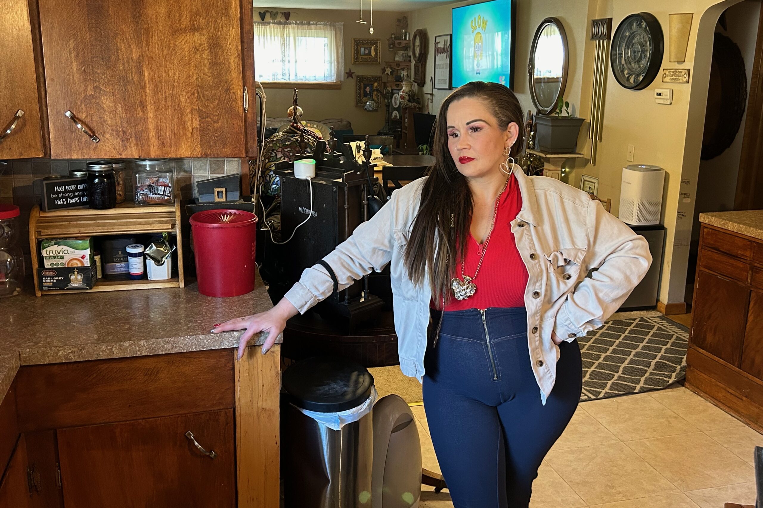 A woman in a red shirt and tan cardigan and jeans stands in her kitchen.