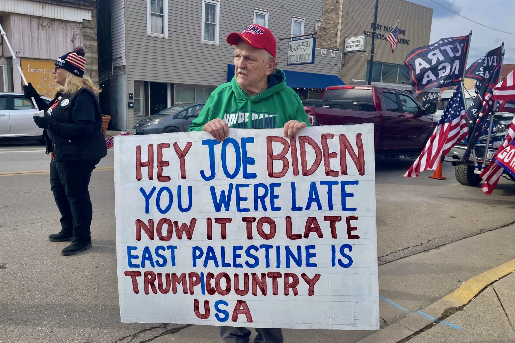 Don Skowron, wearing a red MAGA hat, stands on the street with a sign that reads, "Hey Joe Biden. You were late. Now it's too late. East Palestine is Trump country U.S.A.," in Downtown East Palestine.